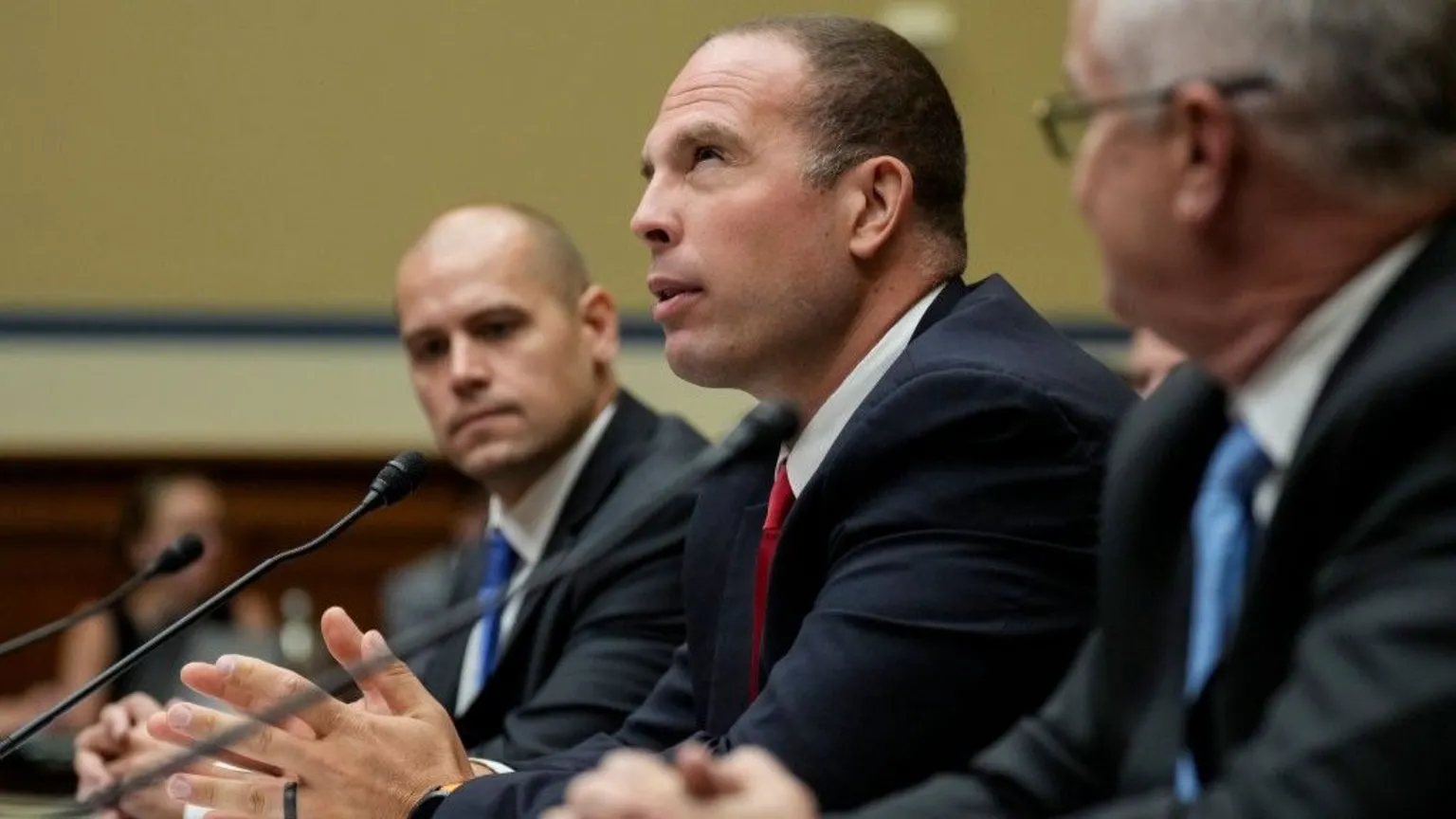 Three men, all wearing dark suits and are seated at microphones.