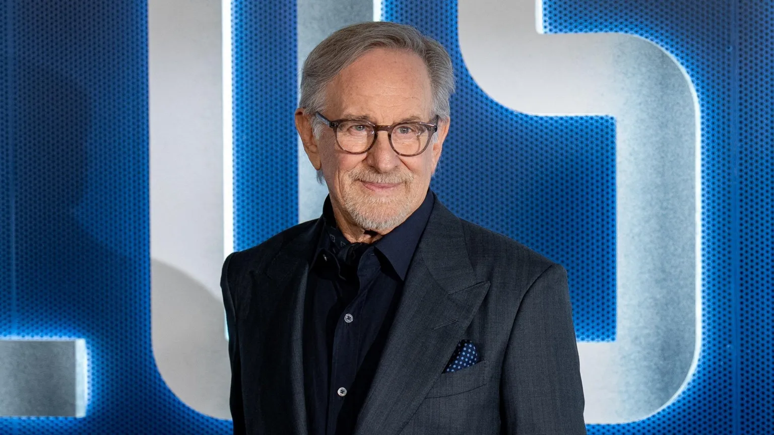 Steven Spielberg looks off camera. He is smiling and wearing a dark grey suit with dark blue open-necked shirt and a spotted blue handkerchief in his breast pocket.