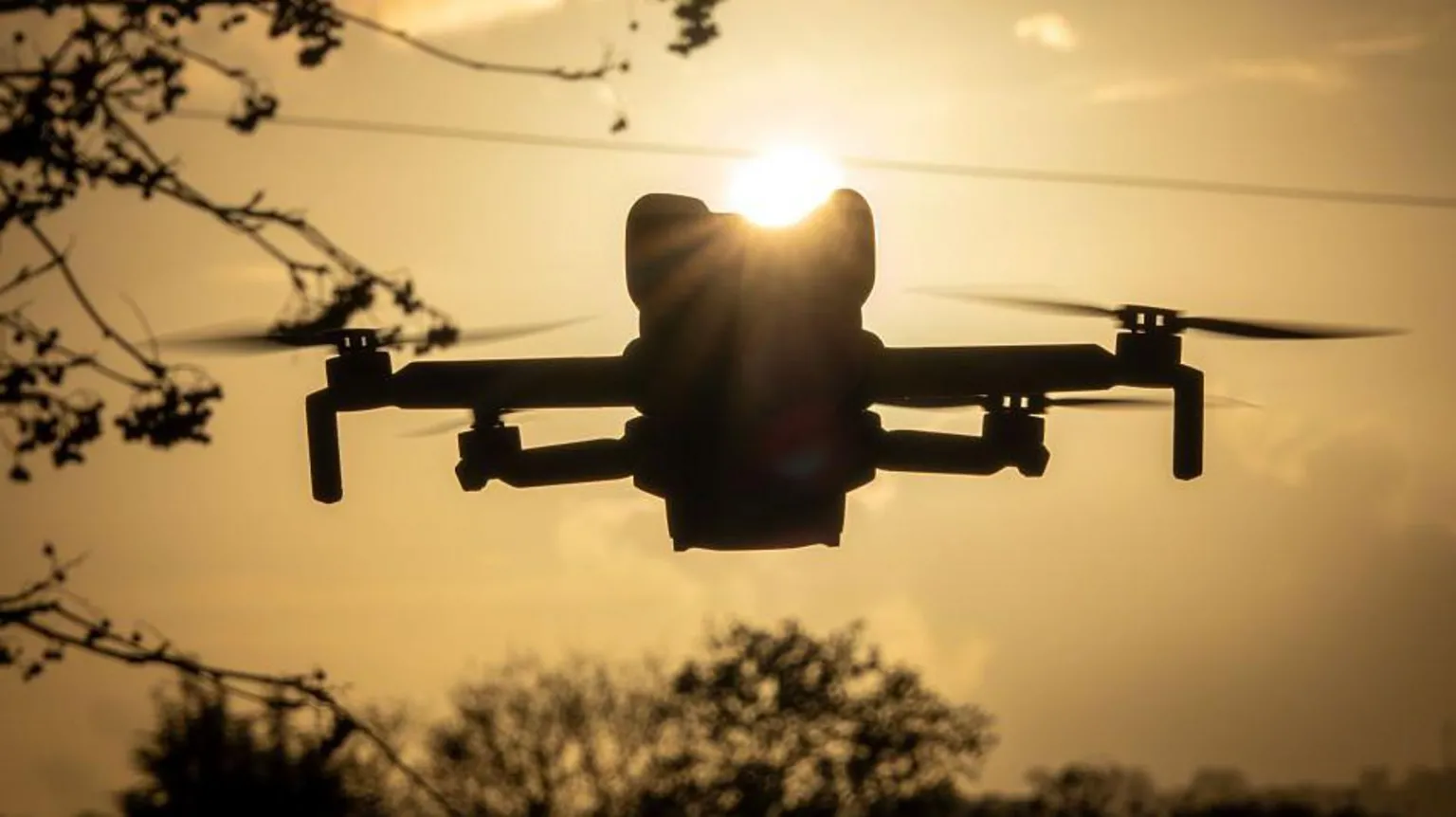 A small drone with four propellers hovers near some trees. It is either near dawn or dusk as it appears in silhouette with the sun behind it.