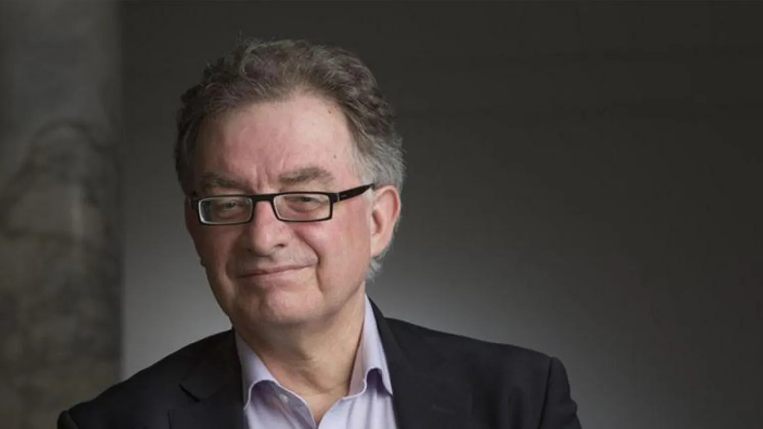 Bill Robinson Prof Chris French looks directly at the camera and smiles. He is wearing a black suit jacket with open-necked light mauve shirt. He has short dark grey hair and rectangular, dark-framed glasses. There is a dark grey background behind him.