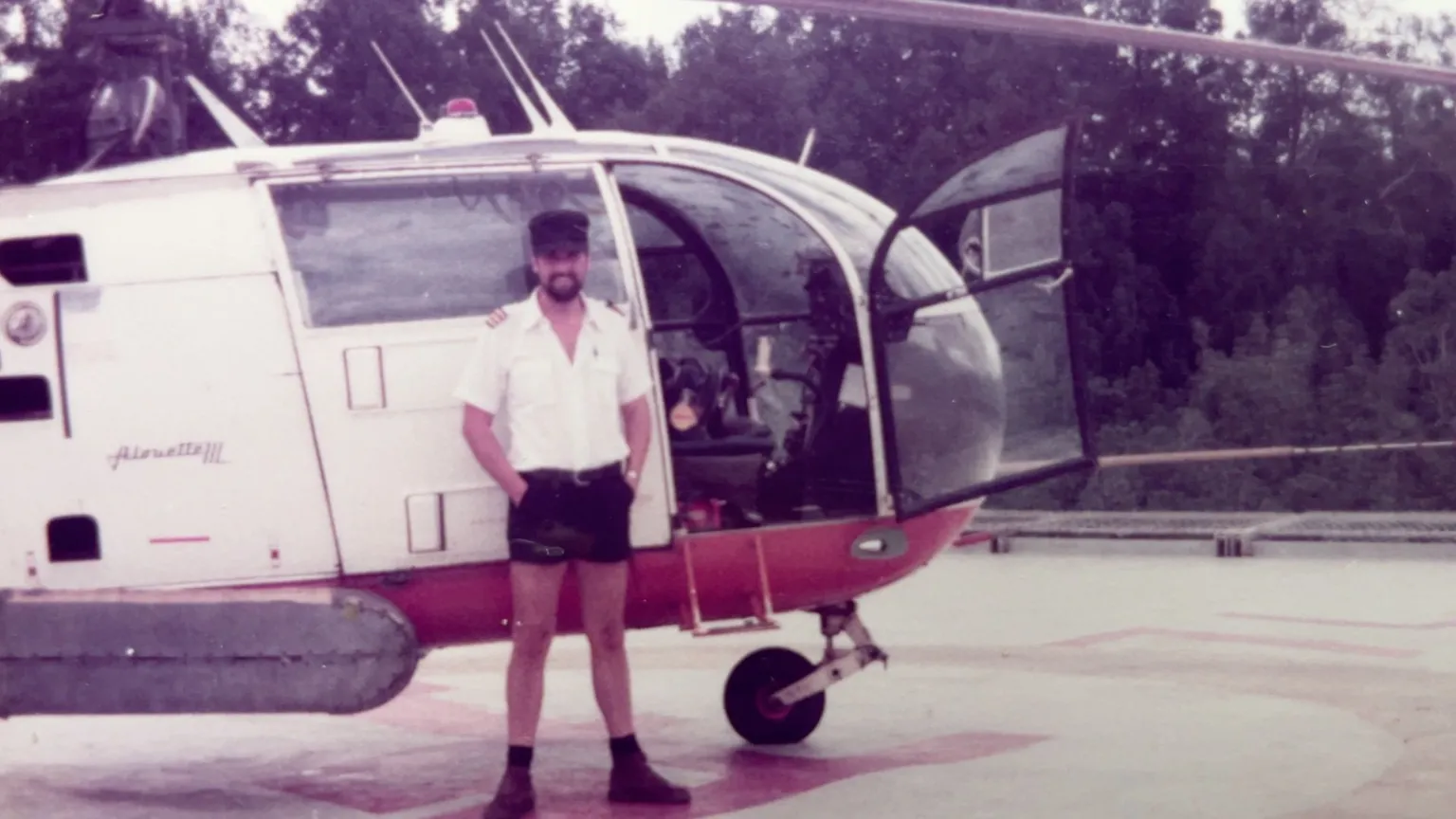 Chris Crowther Chris Crowther, wearing a white shirt and black shorts, stands by a white and red helicopter which has its cockpit door open. He is wearing a black cap.