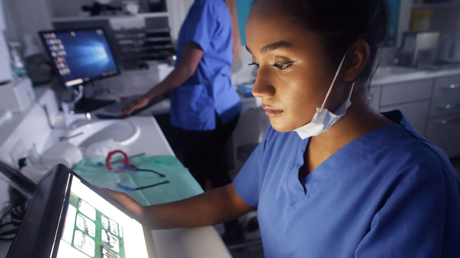  Two health care workers at a clinical station. One is sitting down and looking at a computer screen, which appears to have X-rays on it. Both are wearing blue v-necked tops. The other is in the background, standing up at a computer