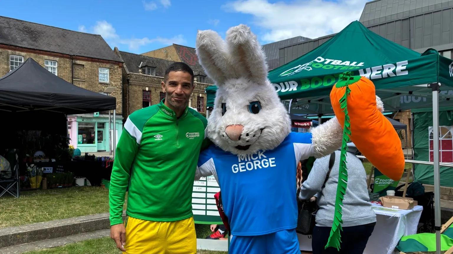 Jenny Kirk/BBC David James wearing a green shirt and yellow trousers standing beside a person dressed as Peterborough United's mascot Peter Burrow. It has a white rabbit head, is holding a large stuffed carrot and is wearing a Peterborough United blue and white strip. Behind are various gazebos with people sitting underneath them. They are in Peterborough city centre.
