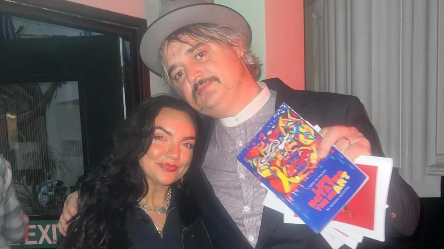 Georgia Griffiths Georgia stands and smiles next to Pete who is holding a copy of her poetry book. Pete, on the right, wears a striped shirt, black suit jacket and a grey hat. He has his arm over Georgia, who wears a denim jumpsuit and has curly long black hair.