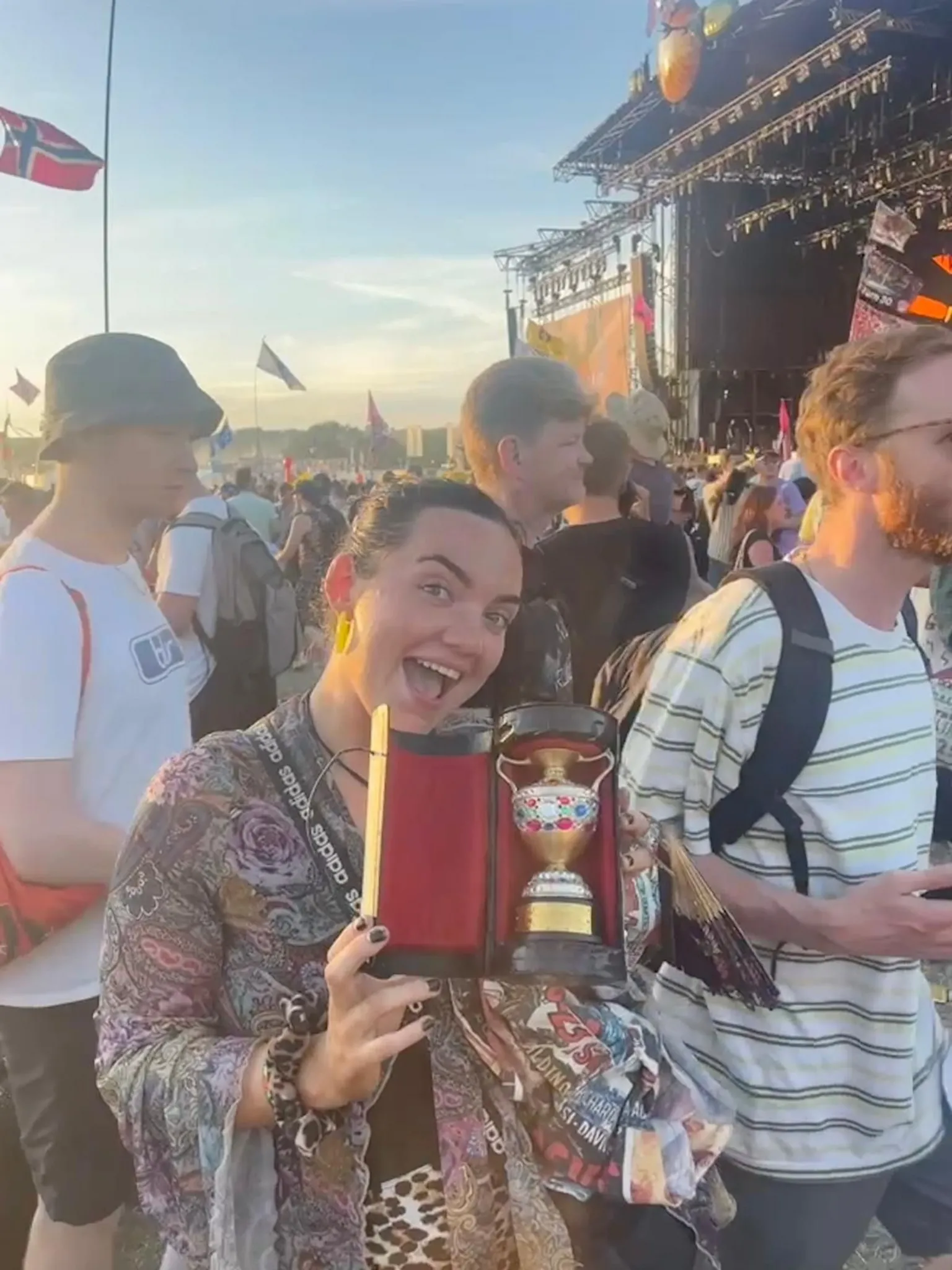 Georgia Griffiths Georgia smiles holding her trophy with crowds behind her at Glastonbury festival. She wears a bohemian blouse, leopard print skirt and has black hair in a ponytail.