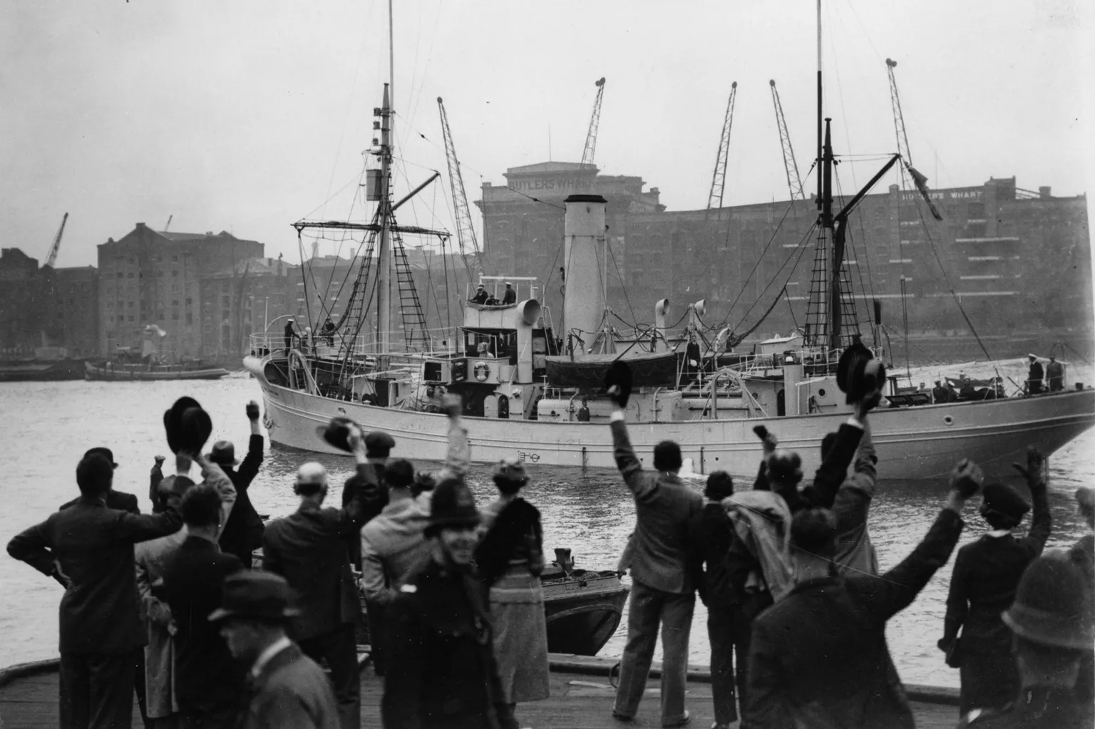 Imagno/ A black and white photo showing a small steam ship sailing across a dock as people in the foreground wave it off. The ship is painted in a light colour and has a single funnel and two masts. People can be seen on the deck. Dock warehouses can be seen in the background.
