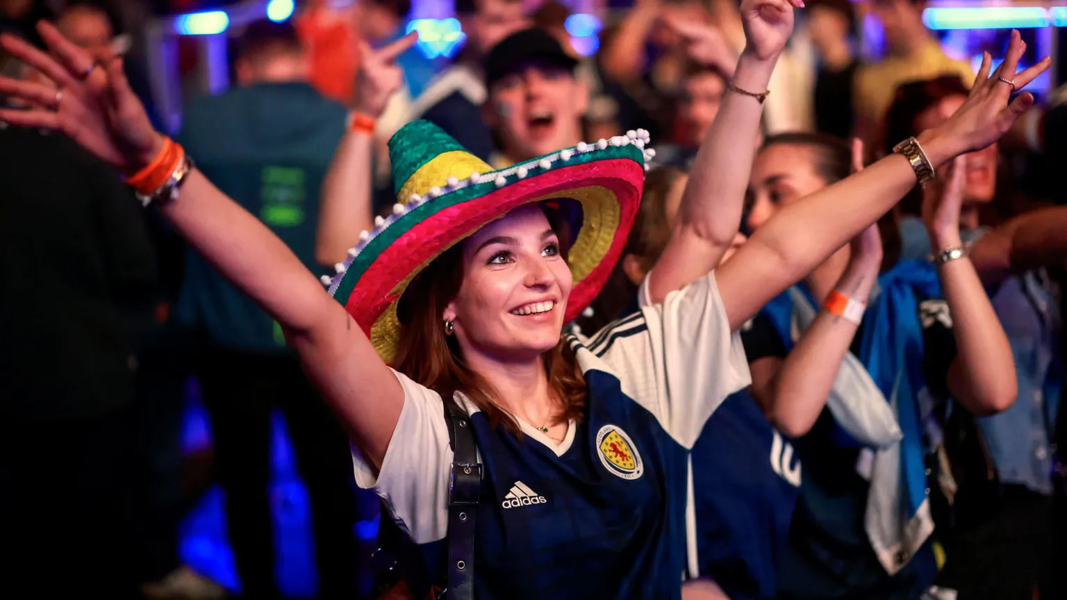 A female fan wearing a colourful hat and a Scotland strip celebrates with her arms outstretched in the OVO Hydro. Rows of supporters can be seen in the background.