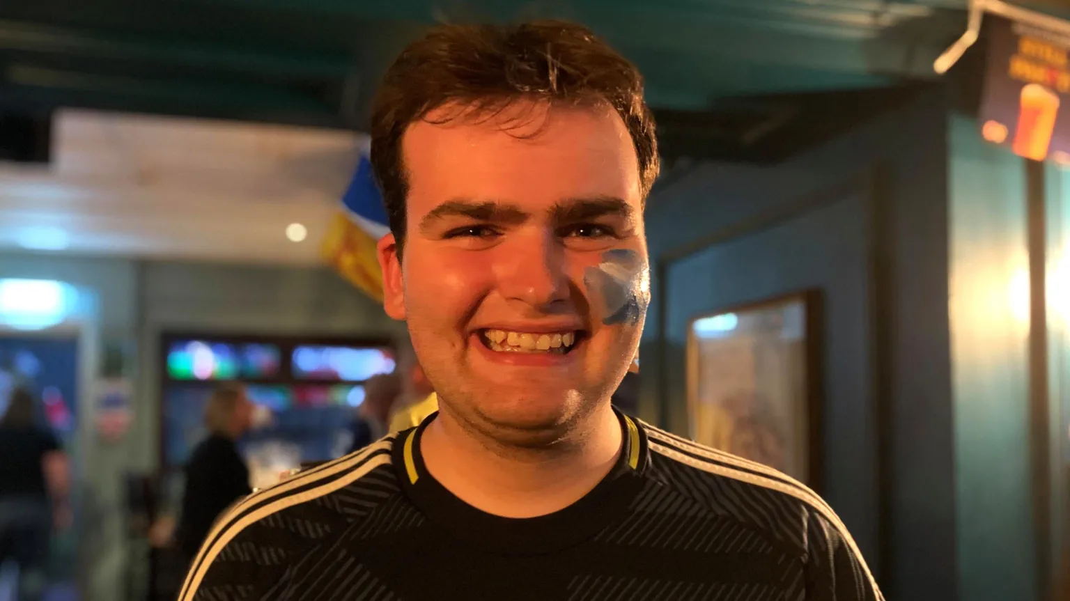 Shaun McGeachan, wearing a Scotland top and with a saltire painted on his left cheek, smiles while standing inside a pub.