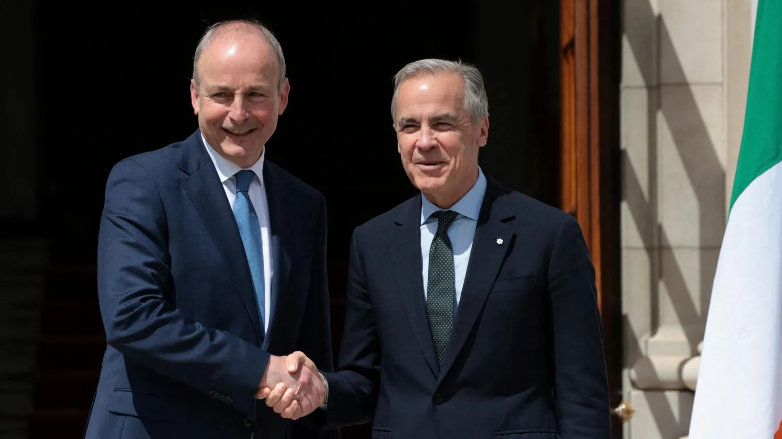  Martin and Carney shake hands and a tri-colour hands to the right hand side of the pair. Martin a bit taller and bald on top. Carney has short grey hair. 