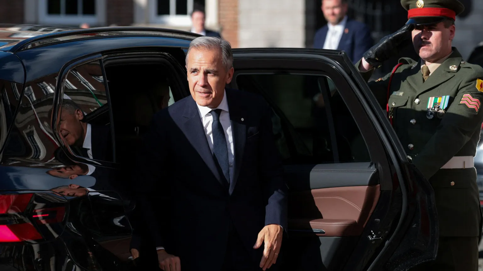  Carney steps out of a shiny black car. He wears a sharp suit. Behind him a solider salutes. 