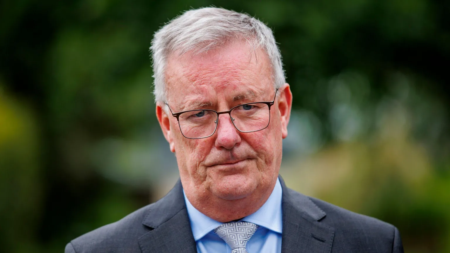 Mike Nesbitt is standing outside with trees behind him. He wears glasses and has a grey suit on and a blue shirt and is wearing a tie with a maze pattern.