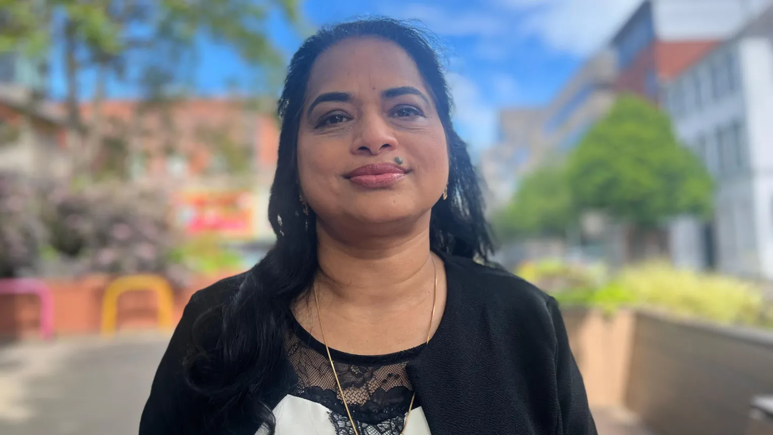 Biji Jose is standing in a street and looking into the camera. She's wearing a black jacket and her top is black and white with lace.