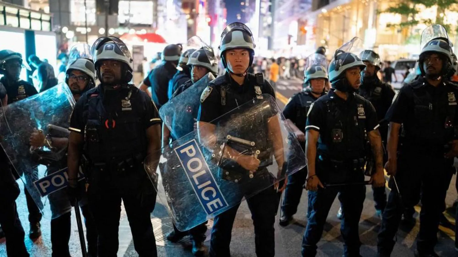 A group of police officers wearing helmets and holding shields prepare to disperse a crowd in Bryant Park, New York City.