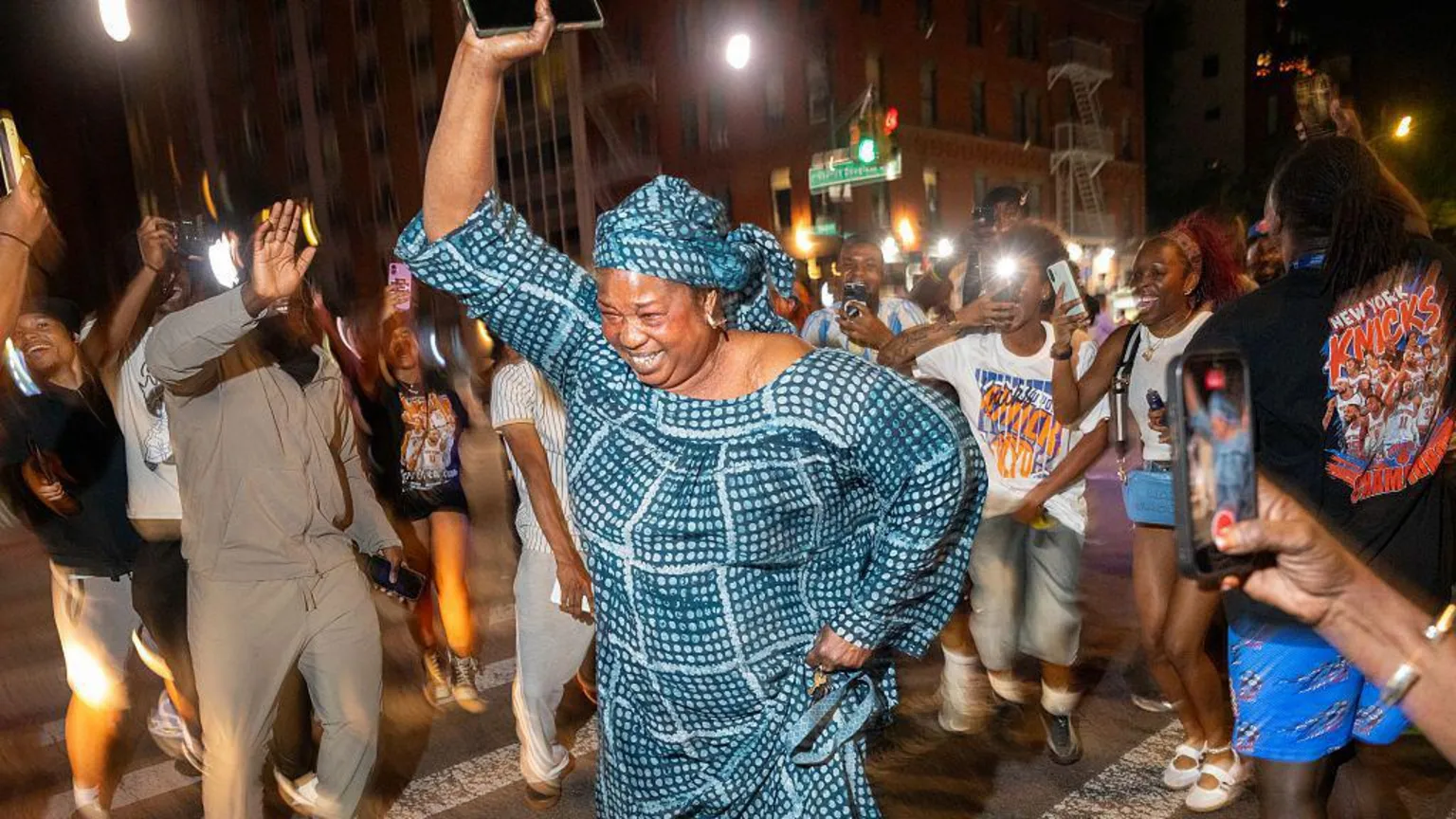 A woman wearing a matching headscarf and dress raises her arm in the air as she dances on a zebra crossing in the street at night time. A crowd of other people dance around her, some raising their phones taking pictures.