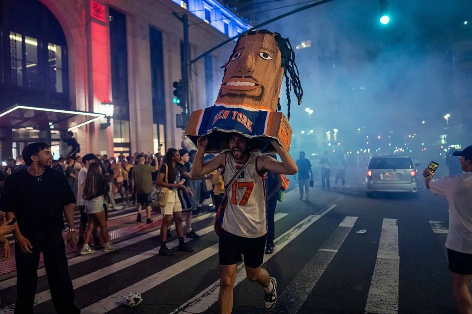 A fan runs in the street holding a cardboard sculpture of a New York Knicks player's head and torso.