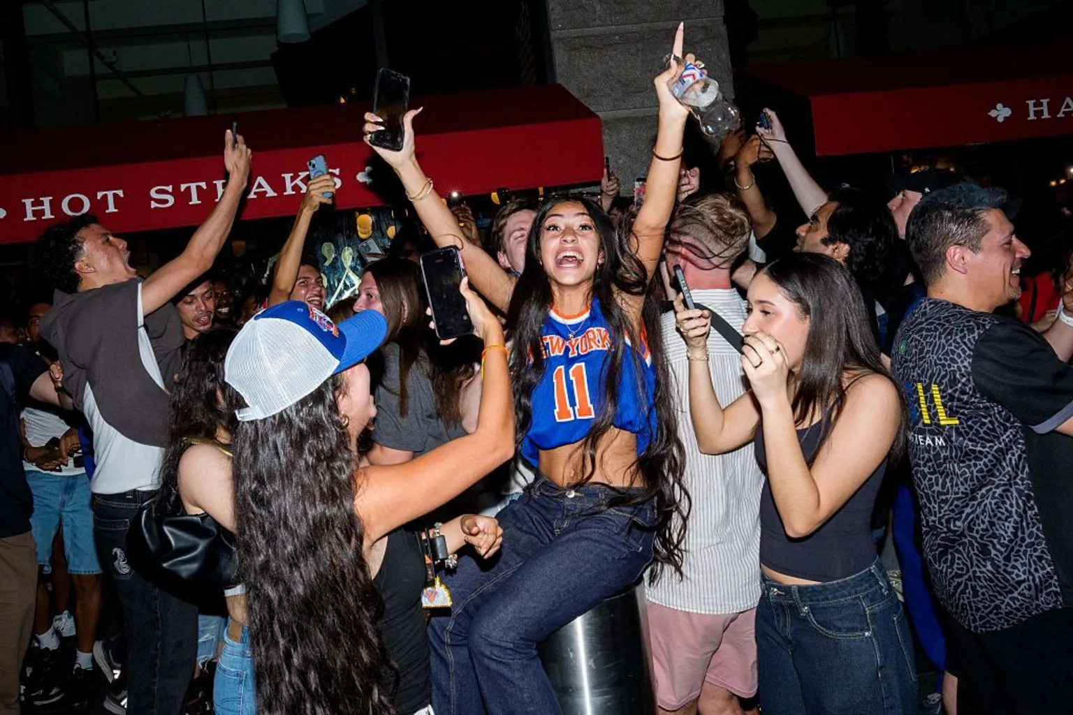 A young woman wearing a Knicks jersey raises her hands in the air in celebration, surrounded by other young people.