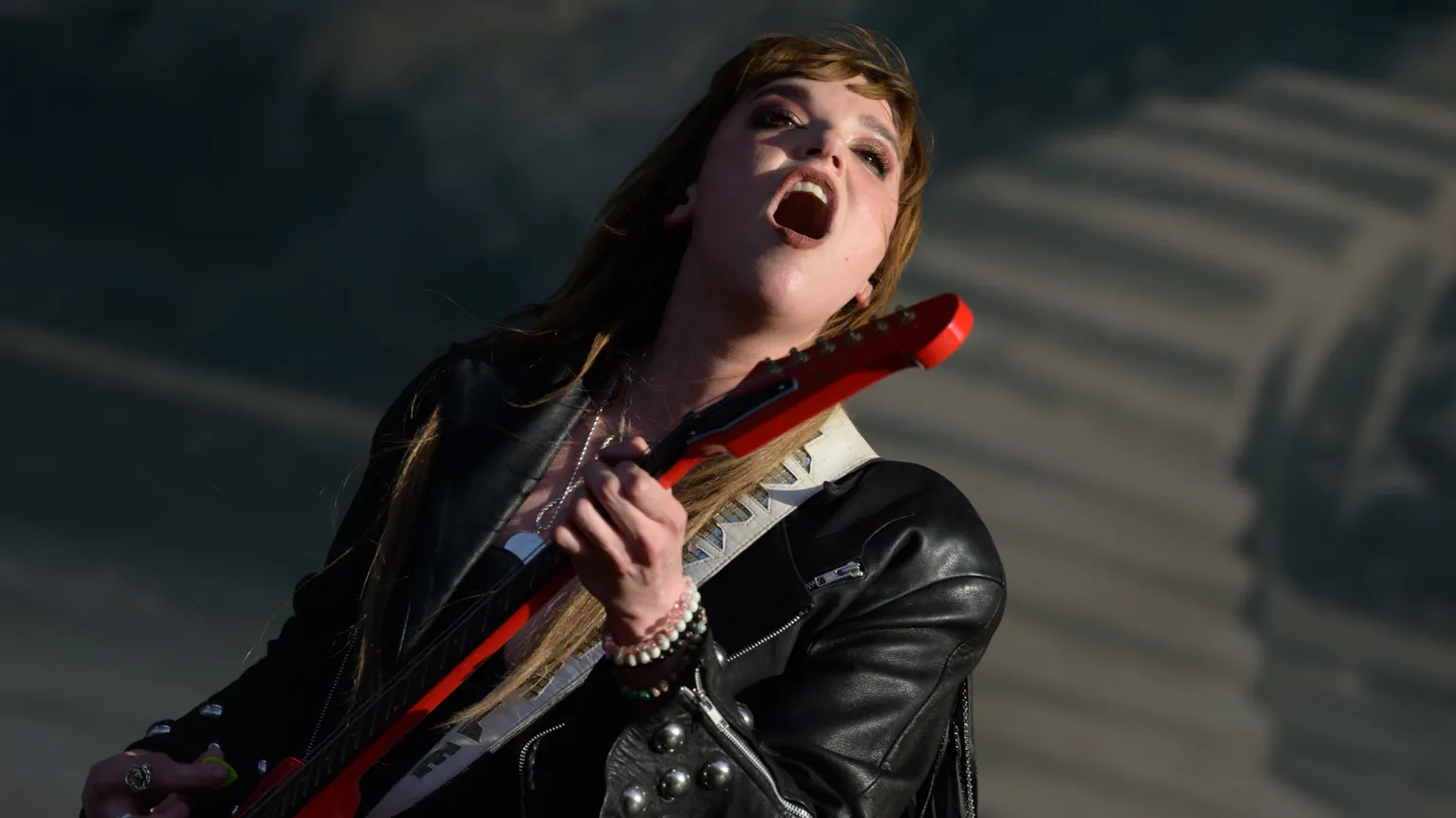  A woman singing on stage. She has long brown hair and is wearing a black leather jacket and holding a red guitar