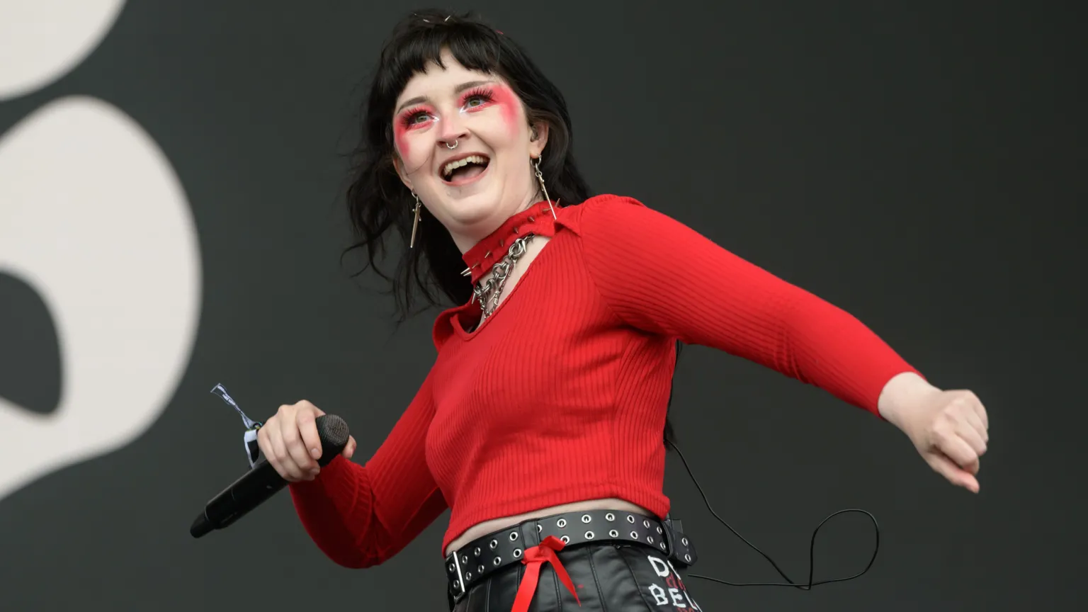  A woman performing on stage. She has black hair and is wearing red face paint and a red jumper. He is holding a microphone. She looks like she is dancing and is smiling.