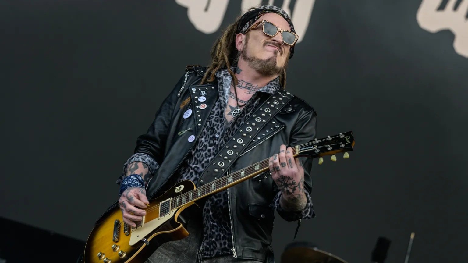  A man stands on a stage holding a guitar. He has dreadlocked hair and is wearing a bandana, sunglasses, a black leather jacket with metal studs, and a leopard-print shirt. He has tattoos on his hands, arms and neck