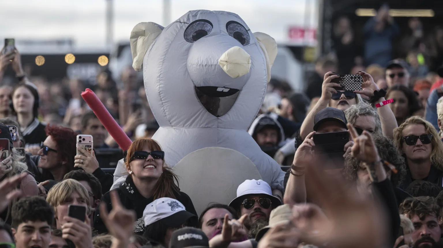  A person in a large, white rat costume stands in the middle of a crowd of people, many of whom are holding up their mobile phones