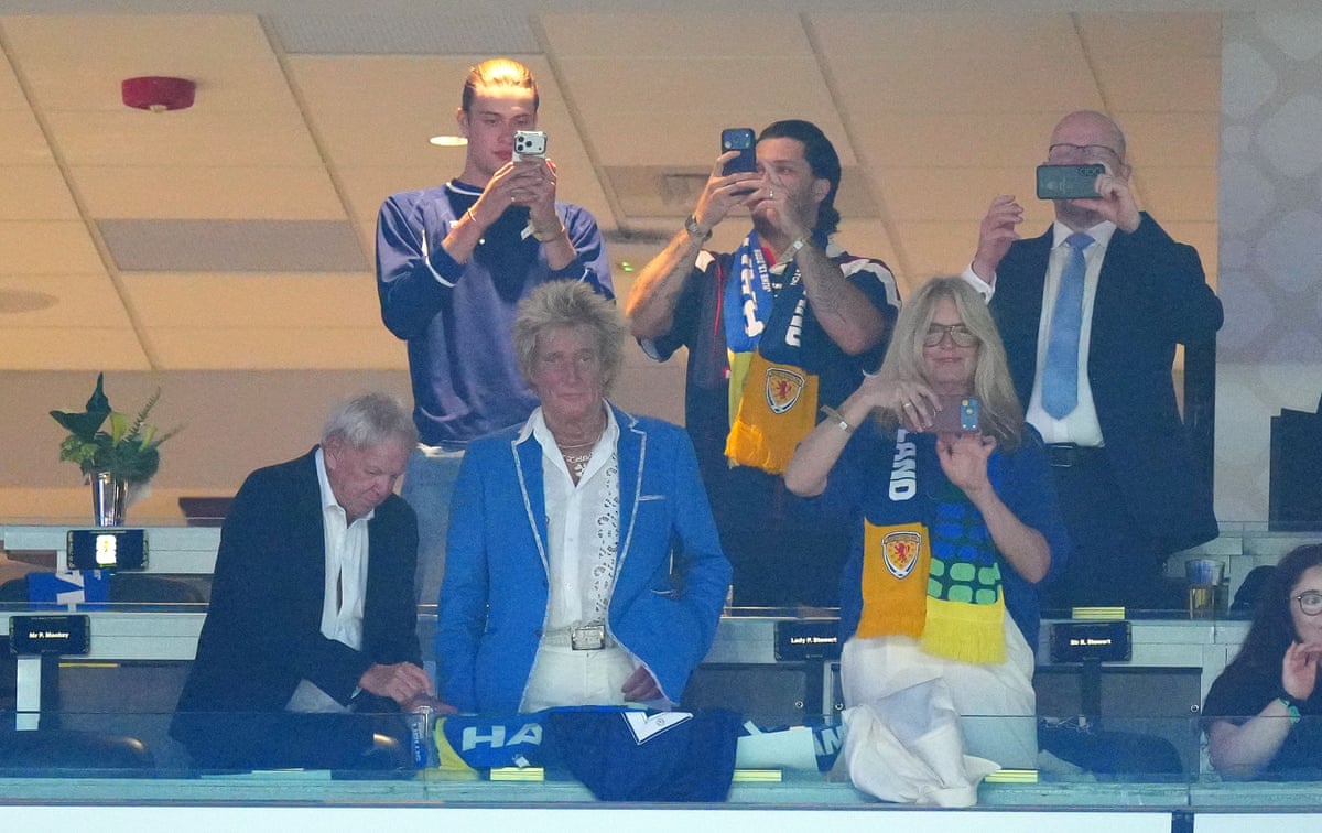 Stewart in a blue blazer watching from a box as people take photos on phones