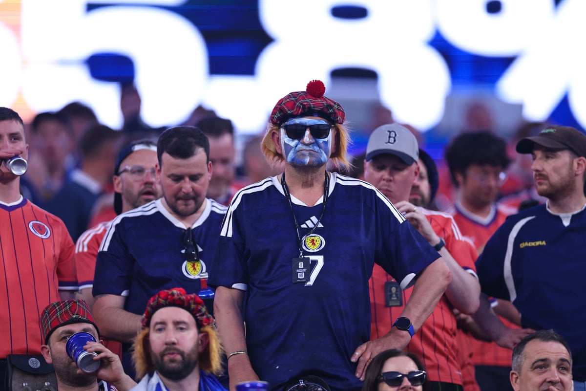 A man standing in the crowd in a tartan hat with his face painted like the Scotland flag