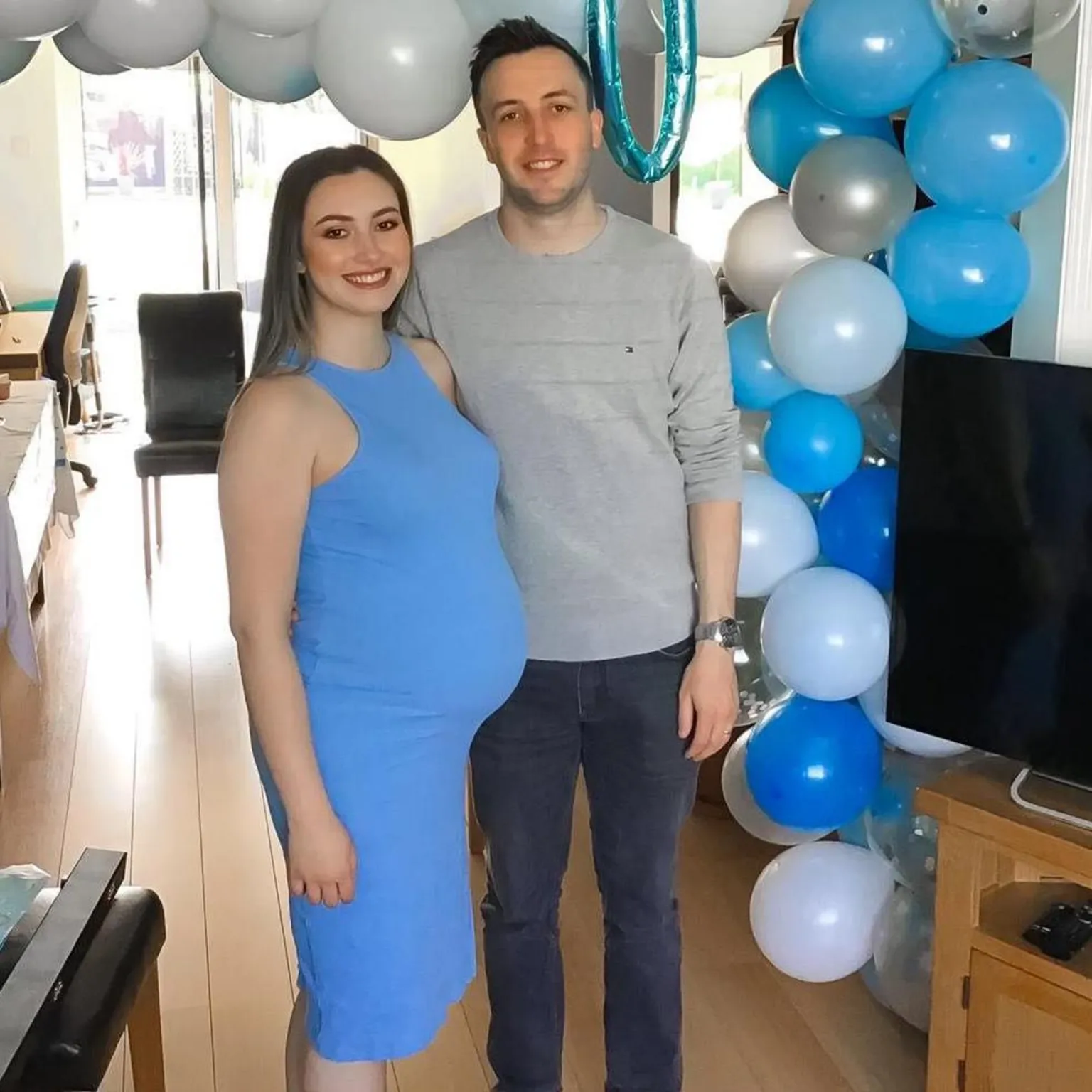Kianty and Conor Simpson Kianty in a blue dress with a visible baby bump is smiling next to Conor. There's a table of buffet food in the background and a balloon arch with white and pale and dark blue balloons. A metallic blue balloon at the top of the arch reads ''Baby