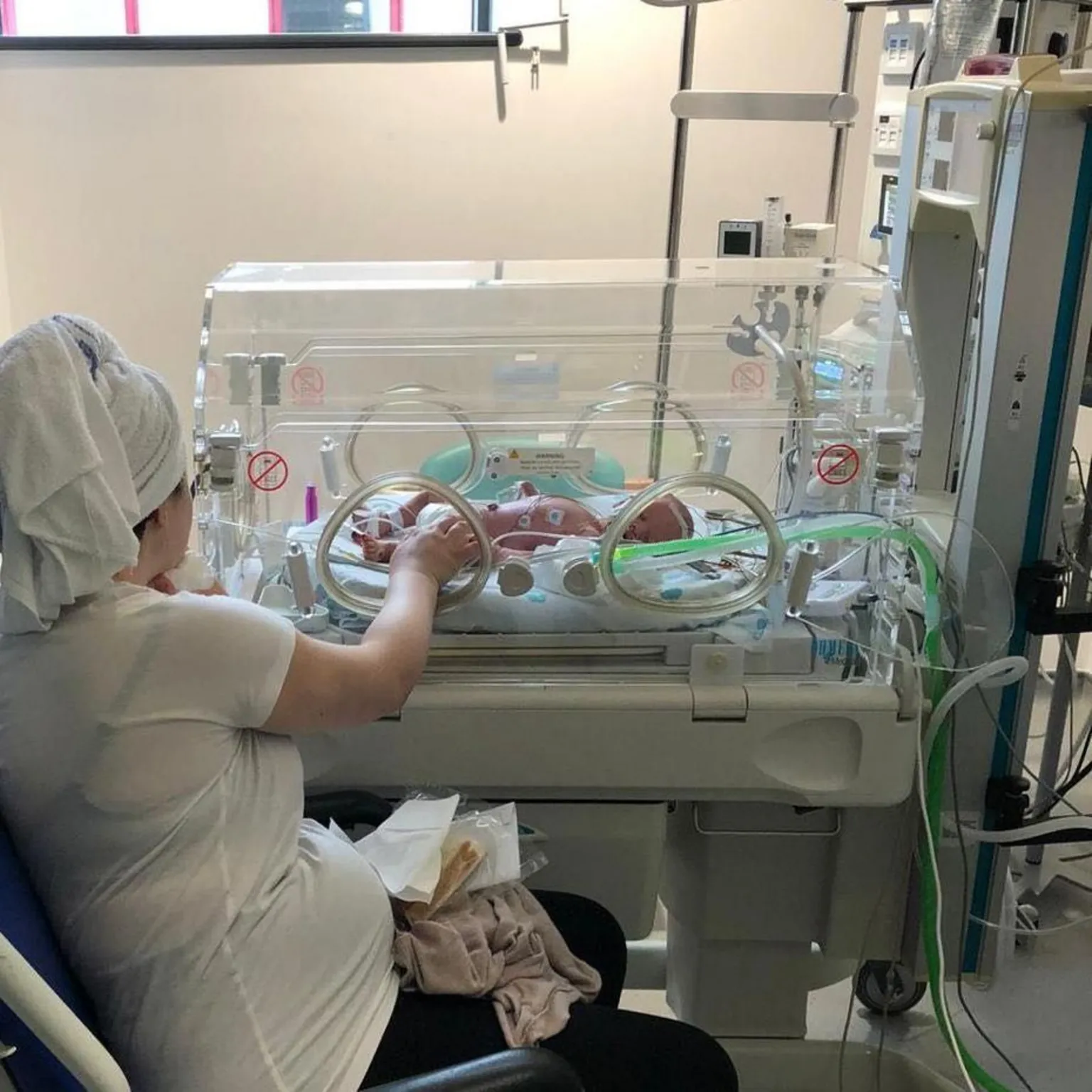 Kianty and Conor Simpson Kianty with her hair wrapped up in a towel sits in a hospital wheelchair in front of an incubator. She's reaching through a circular window in the incubator to touch her baby son's hand.