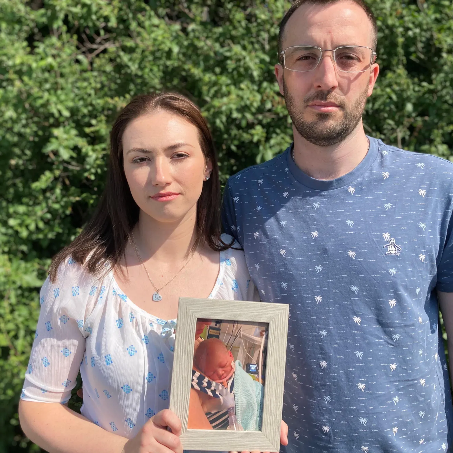 Kianty and Conor face the camera, with Kianty holding a framed photo of Jacob. Both have a serious expression. Kianty has long dark hair and wears a white top with a blue diamond pattern on. Conor has glasses and a short dark beard, and wears a blue top with palm trees on.