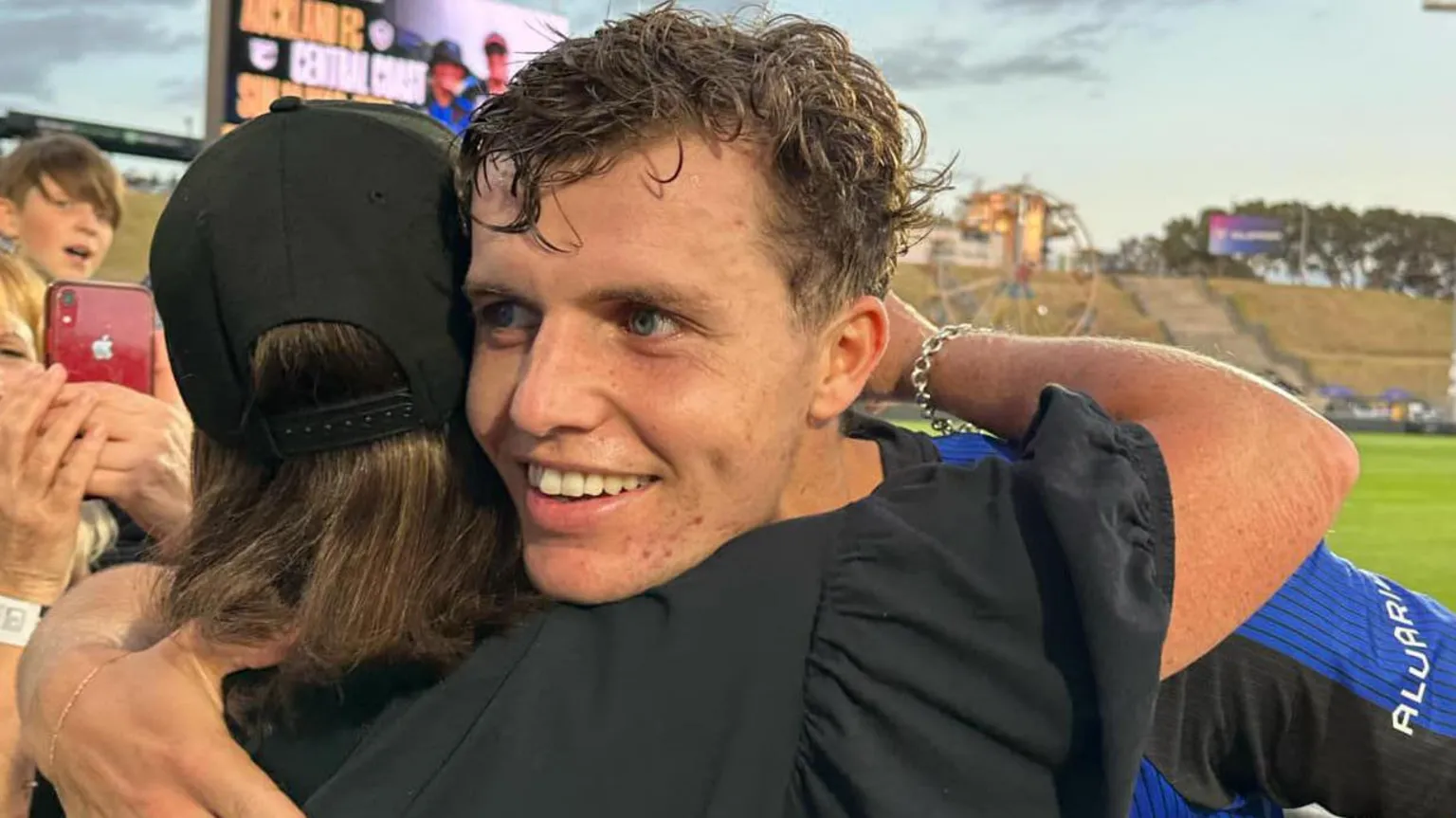 Steve Elliot A woman in a black cap with brown hair and a black top has her back to the camera and is hugging a young man with curly brown hair who looks like he has just finished a game of football.