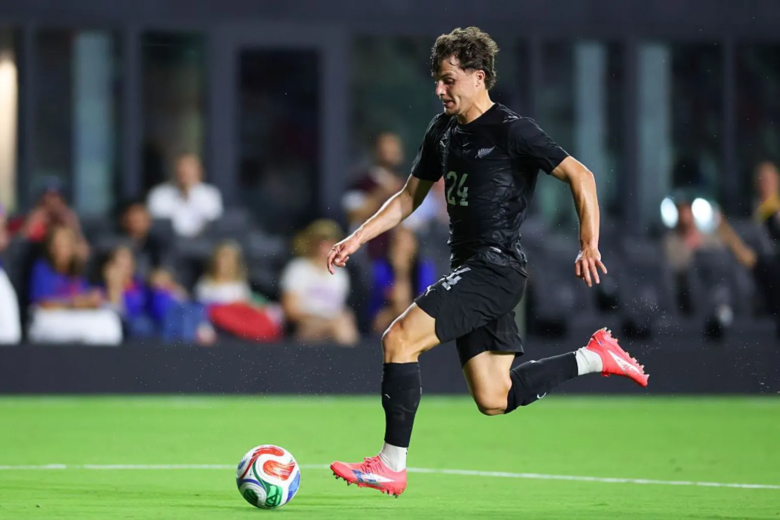 Callan Elliot in a black top, shorts and socks in bright pink boots runs forward with the ball at his feet