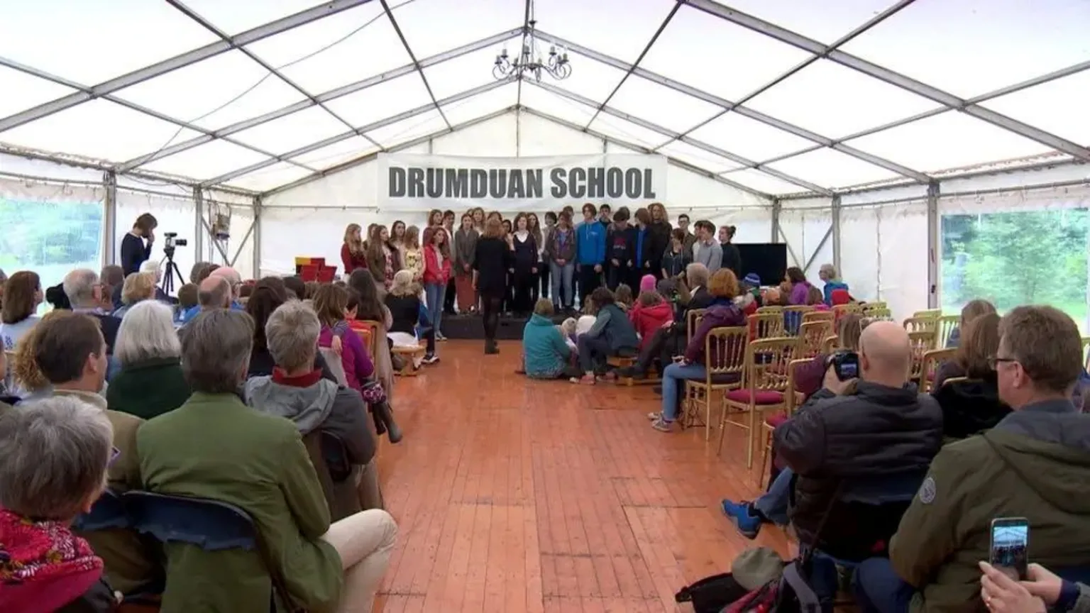 People inside what appears to be a structured tent at a graduation ceremony, with the words DRUMDUAN SCHOOL on a banner.