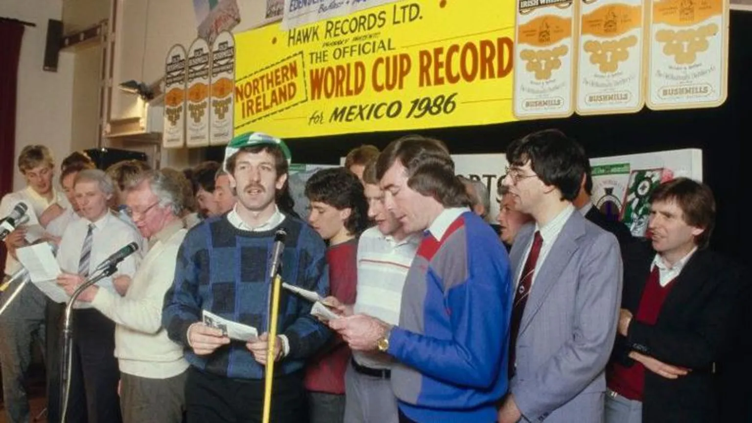 Northern Ireland football team members, including Gerry Armstrong in green and white cap dark hair and moustache and checked jumper. Manager Billy Bingham is in a white jumper and glasses. Pat Jennings is in blue, grey and red jumper.