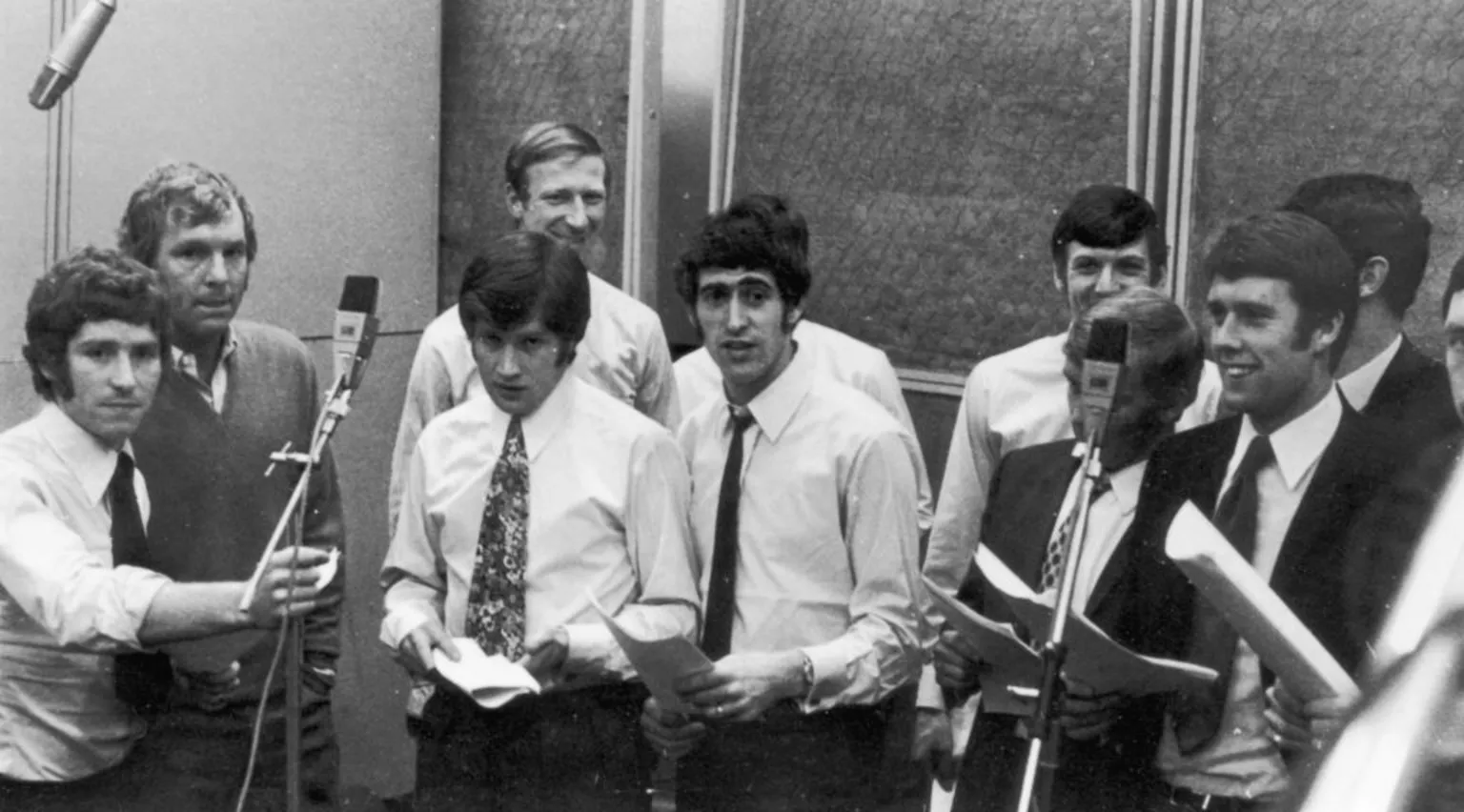 A black and white image of the England World Cup squad ahead of the 1970 tournament. They are in a recording studio with microphones and are holding sheet with lyrics written on them