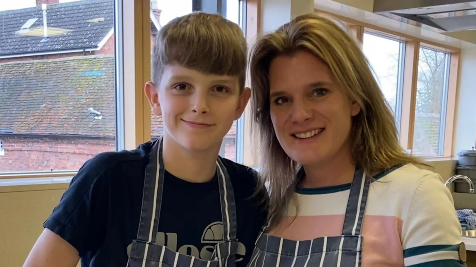 Ellen Roome and her son Jools Sweeney stand side by side in a large kitchen. They are both wearing dark blue aprons with thin white stripes on them.
