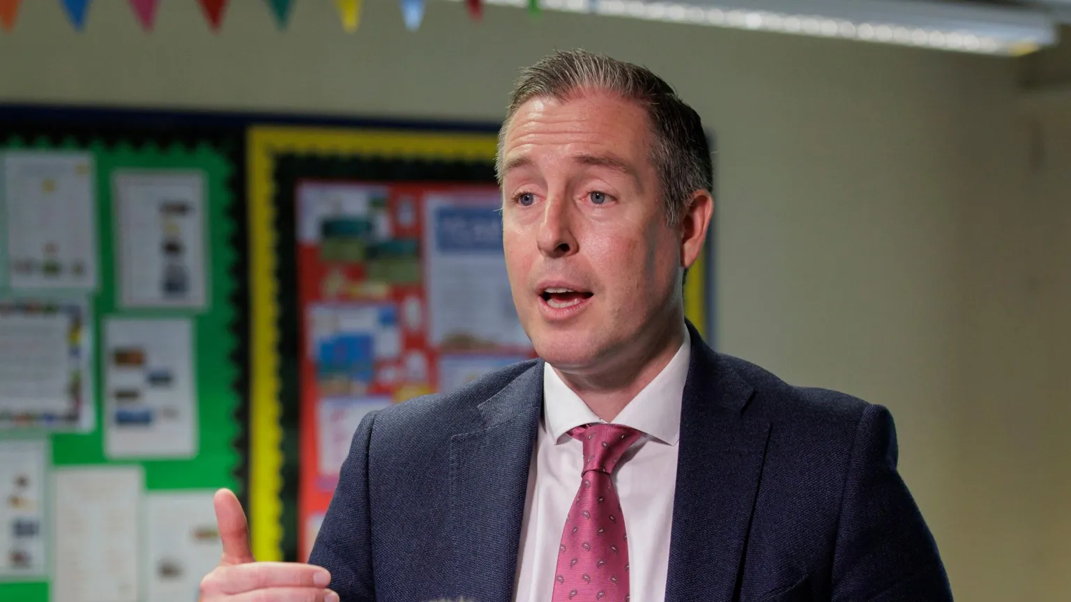 Paul Givan. He has short grey-ish hair, wearing a navy blazer, white shirt and pink tie. Classroom displays are bring him.