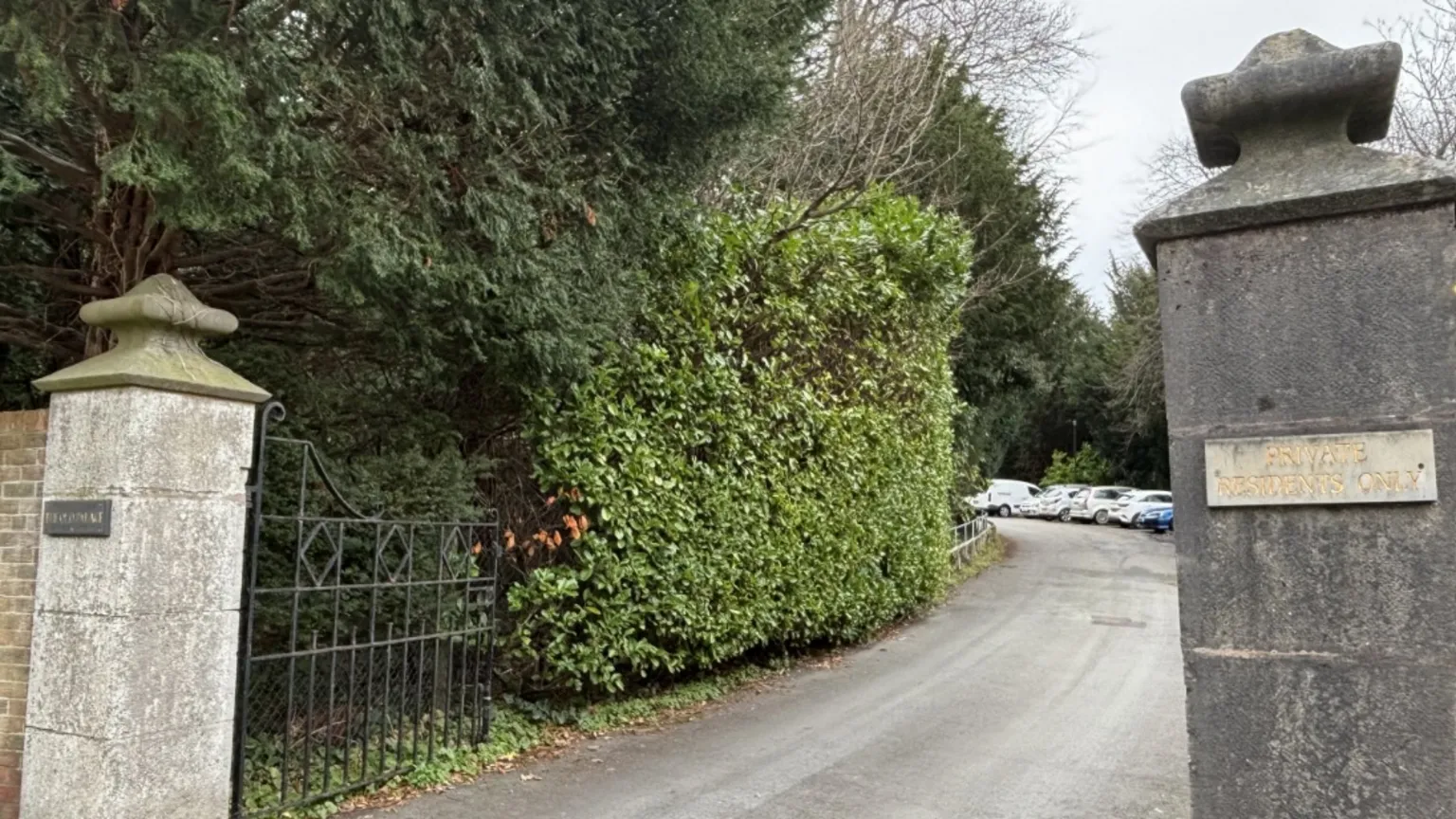 Two pillars on either side of a long driveway with green hedge along one side and a black gate swung open.