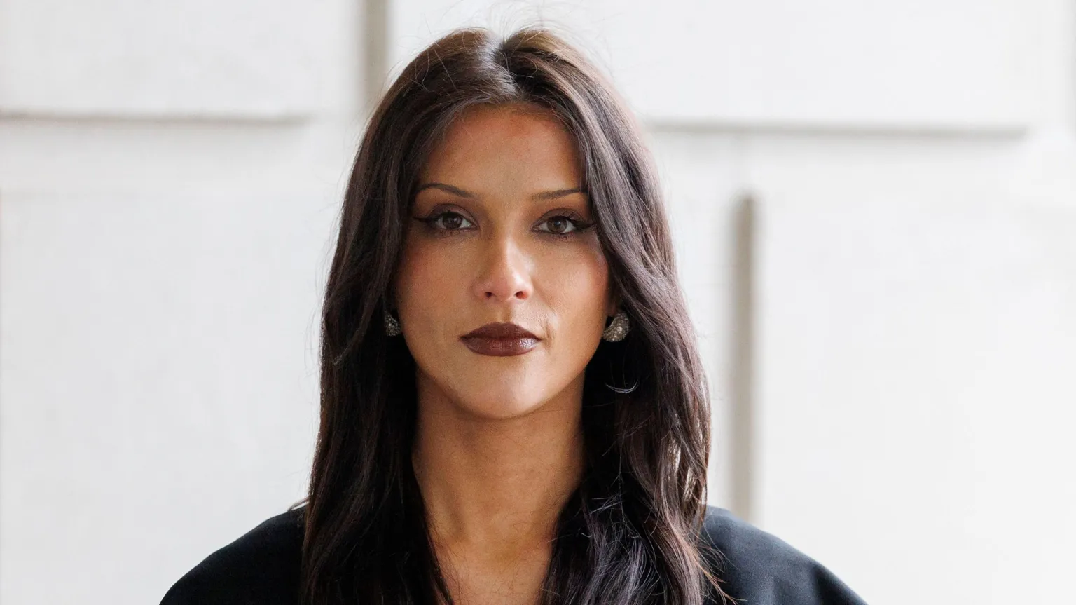 Cara Hunter has long dark hair and brown eyes. She is wearing a black top, silver earrings and dark brown lipstick.