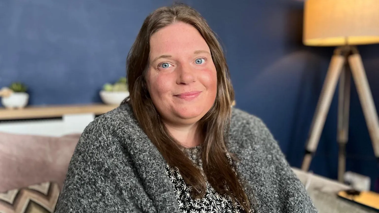 Louise is sat on a sofa in her living room. She has long, dark hair and is smiling at the camera. She is wearing a black and white patterned dress with a grey cardigan.