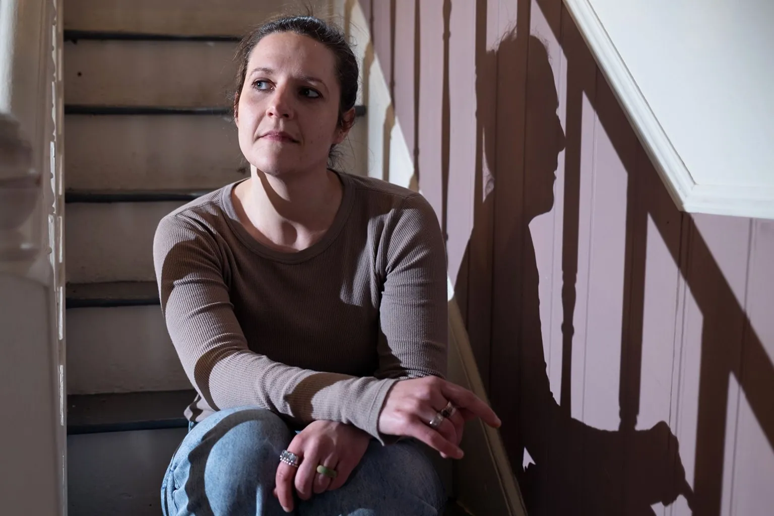 Ruth is sitting on an indoor staircase, illuminated by side lighting that casts a clear shadow on a paneled wall, looking slightly to the side with one arm resting on a raised knee and wearing a long-sleeved top and jeans.