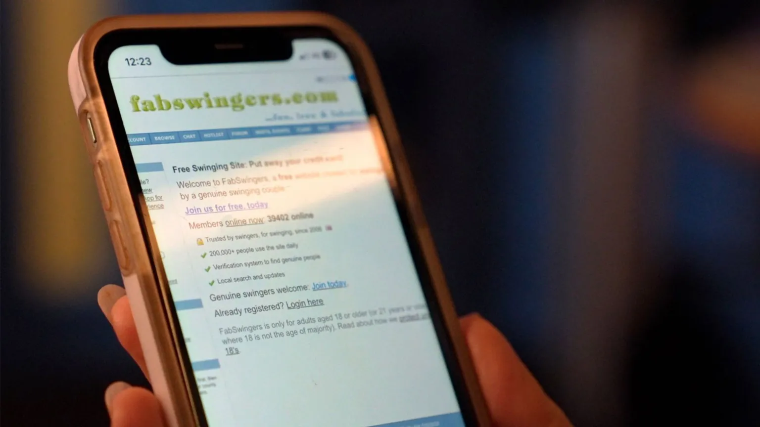 Close-up of a smartphone displaying the FabSwingers website homepage, held in a hand, with the screen slightly out of focus against a dimly lit background.