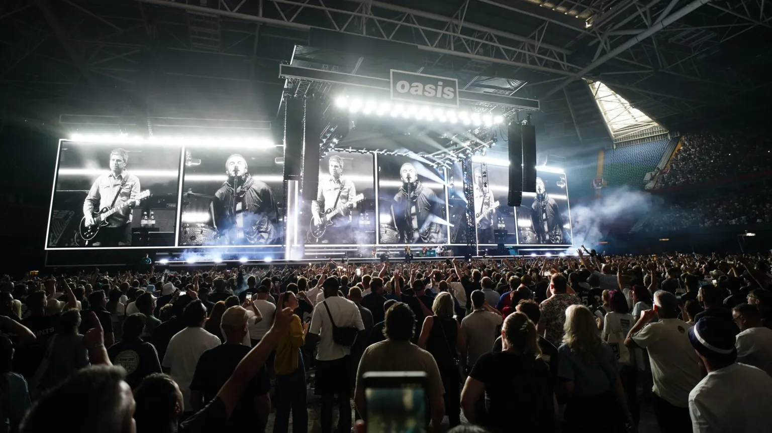 Crowds at the Oasis concert standing and facing the stage. A screen behind the stage shows the Gallagher brothers in black and white, Noel playing a guitar and Liam singing. The Oasis band logo can be seen at the top of the stage.