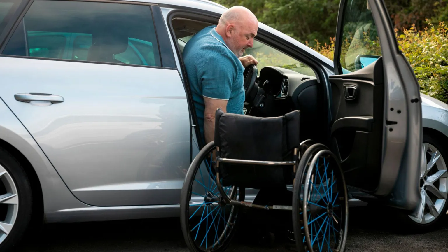 A man is in the middle of getting from a wheelchair into the driver's seat of a car - we can see the driver's side door open, where the wheelchair is sitting empty. The man is mostly in the seat with some of his head still leaning out the door. The car is a silver estate, and behind where it is parked is a green bush.