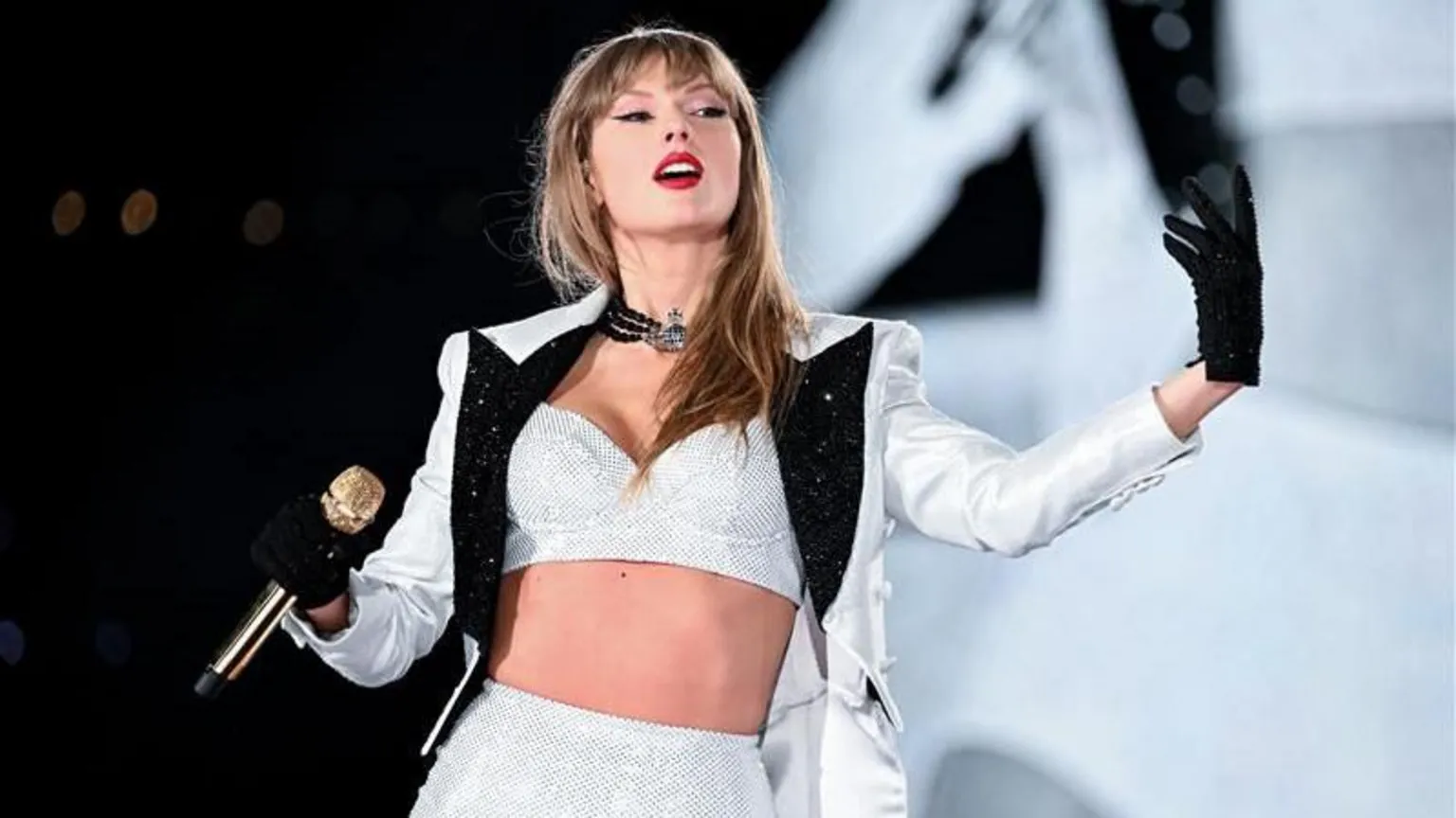 Taylor Swift wearing a white jacket and black gloves. She is holding a gold microphone.