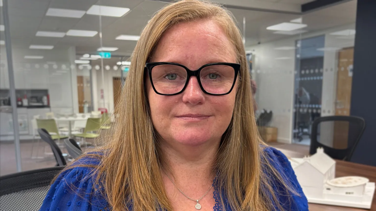Close-up of a person wearing black-framed glasses and a blue top, seated at a desk in a modern office with glass walls and chairs in the background.