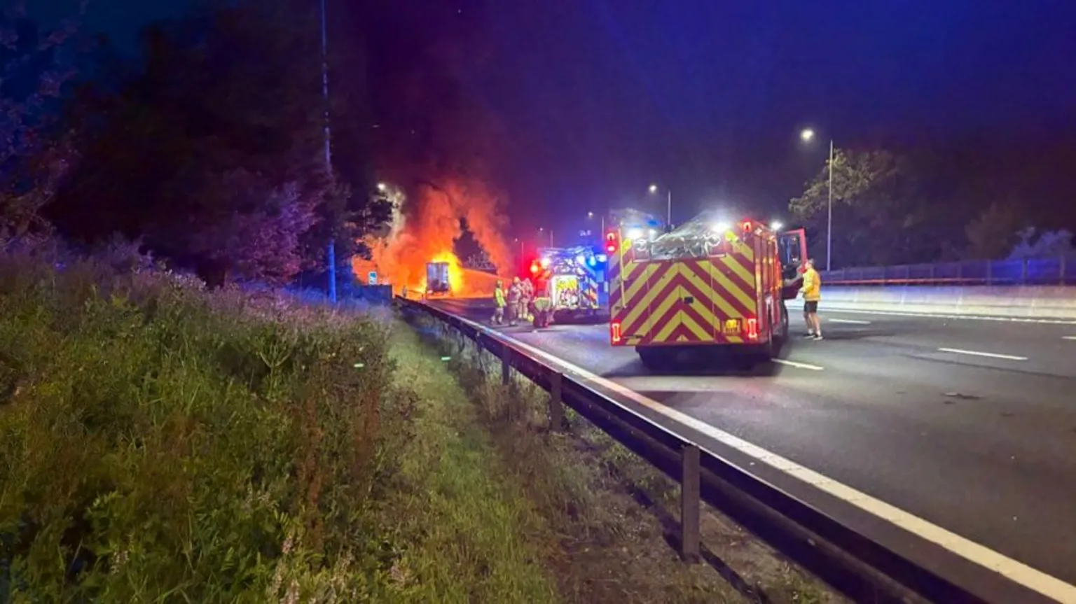 National Highways Two fire engines on the M15, with firefighters are the scene. There is a grass bank and trees to the left. Flames can be seen in the distance.