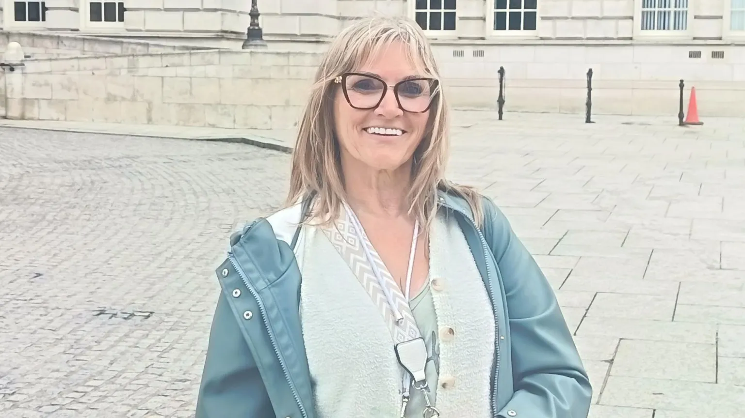A middle aged woman with long grey/blond hair and glasses stands in the grounds of Belfast City Hall