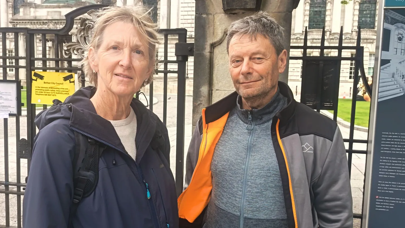 A grey haired woman and man wearing outdoor wear and backpacks stand in front of the gates of Belfast City Hall