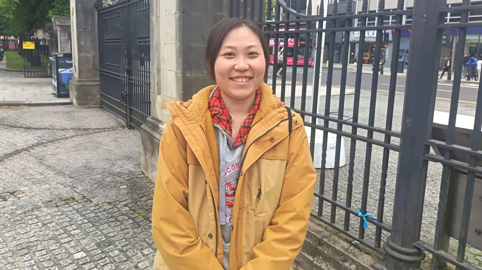 Skylar is an Asian woman with black hair tied back. She is wearing a yellow jacket and is smiling while standing in the grounds of Belfast City Hall