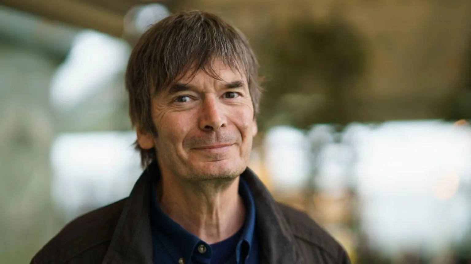 Ian Rankin. He has brown hair and dark eyes and is wearing a brown jacket and blue shirt. The background is blurred.